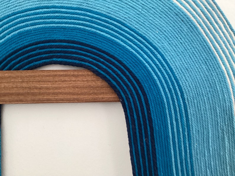 Puede incluir: Una gran pared colgante de hilo azul y blanco en forma de arco. El hilo est&aacute; tejido en un patr&oacute;n degradado, comenzando con azul oscuro en la parte inferior y desvaneci&eacute;ndose a blanco en la parte superior. La pared colgante est&aacute; unida a una varilla de madera.
