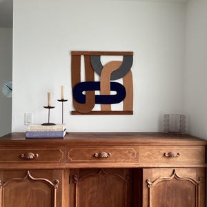 May include: A wooden wall art piece with geometric shapes in brown, gray, and navy blue hangs above a wooden cabinet. Two black candle holders with lit candles and a stack of books are on the cabinet. A glass block window is on the right.