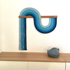 Puede incluir: Un tapiz de pared azul y blanco hecho de hilo, con una barra de madera, en forma de ola. El tapiz de pared est&aacute; montado en una pared blanca sobre un estante de madera clara con una figurita de p&aacute;jaro de cer&aacute;mica azul en el estante.