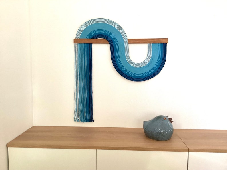 Puede incluir: Un tapiz de pared azul y blanco hecho de hilo, con una barra de madera, en forma de ola. El tapiz de pared est&aacute; montado en una pared blanca sobre un estante de madera clara con una figurita de p&aacute;jaro de cer&aacute;mica azul en el estante.