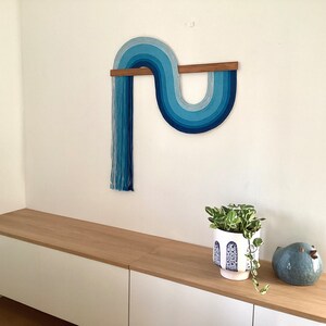 Puede incluir: Un tapiz de pared azul y blanco con una barra de madera, una planta en maceta y una figurita de p&aacute;jaro azul en un estante blanco y de madera.