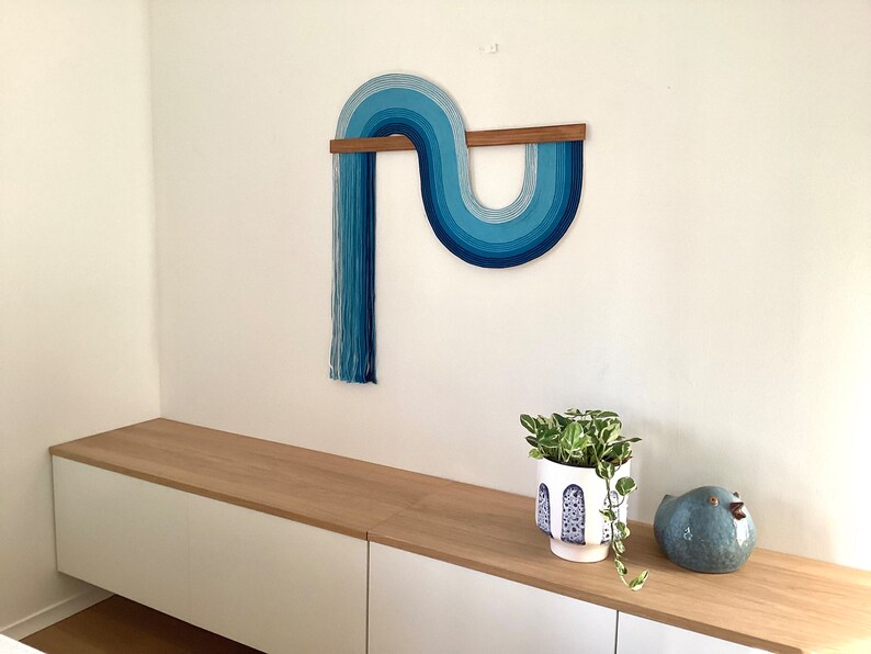 Puede incluir: Un tapiz de pared azul y blanco con una barra de madera, una planta en maceta y una figurita de p&aacute;jaro azul en un estante blanco y de madera.