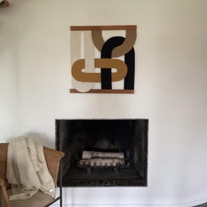 May include: An abstract wall hanging with a wooden frame. The artwork features interlocking shapes in black, tan, and off-white. A wicker chair with a blanket sits in front of a fireplace.
