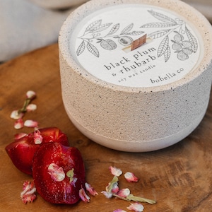 May include: A white ceramic candle jar with a black and white illustration of a branch with leaves and fruit. The candle is labelled "black plum & rhubarb soy wax candle" and "bobella co". The candle is sitting on a wooden tray with red plums and pink flower petals.