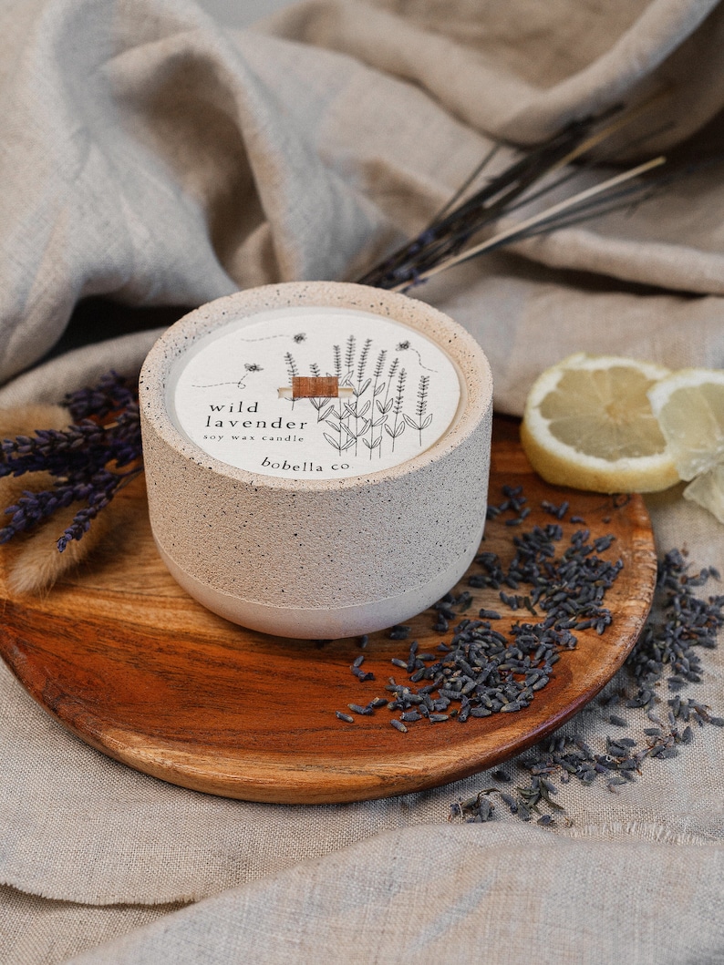 May include: A white ceramic candle with a brown wooden lid. The candle has a label that says "wild lavender soy wax candle bobella co.". The candle is sitting on a wooden tray with dried lavender flowers.