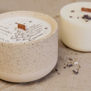May include: A beige concrete candle jar with a white label that says "wild lavender candle ella co." The label has a drawing of lavender flowers and a small wooden tag. The candle jar is sitting on a beige linen surface.