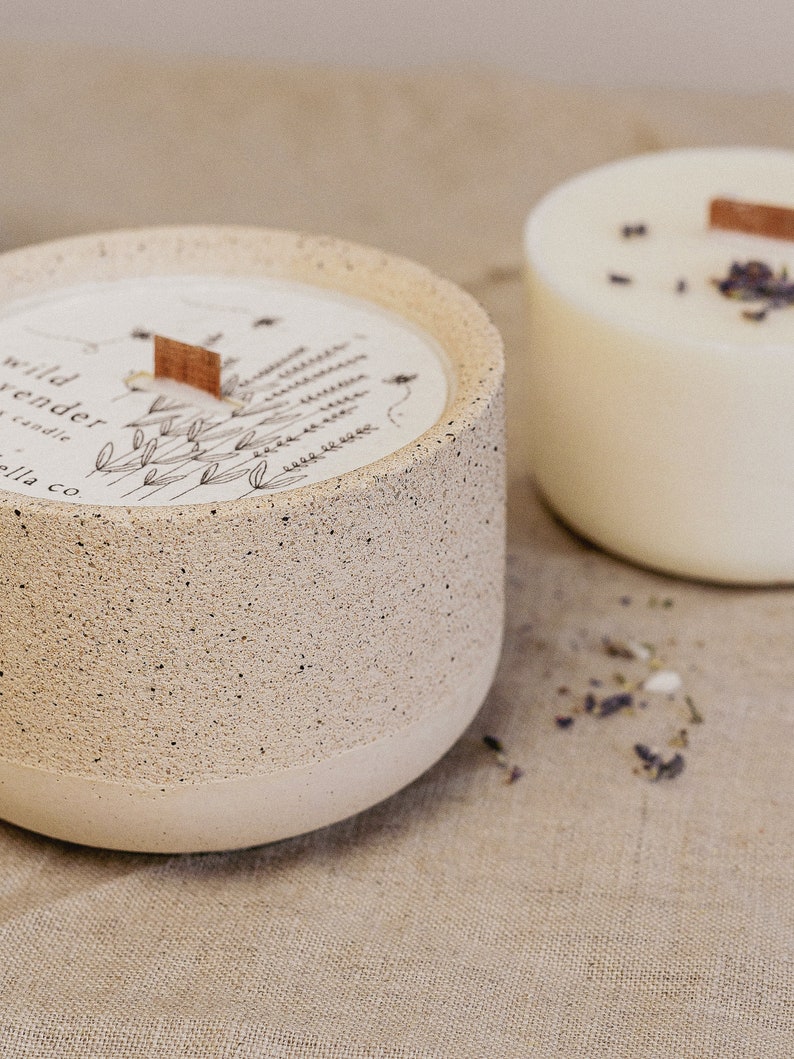 May include: A beige concrete candle jar with a white label that says "wild lavender candle ella co." The label has a drawing of lavender flowers and a small wooden tag. The candle jar is sitting on a beige linen surface.
