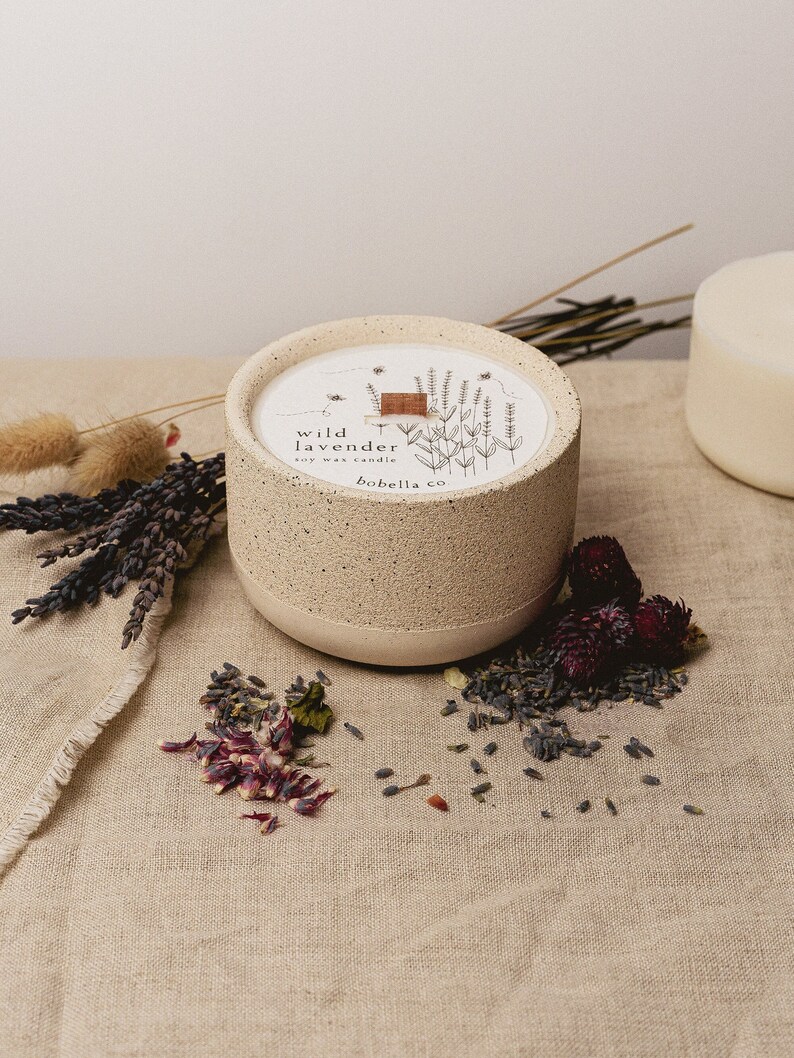 May include: A beige ceramic candle jar with a white label that reads "wild lavender soy wax candle bobella co.". The candle jar is sitting on a beige linen fabric with dried lavender flowers and petals.