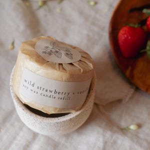 Puede incluir: Recarga de vela de cera de soja envuelta en papel marr&oacute;n, etiquetada como "wild strawberry & vanilla". La vela se encuentra en un soporte de cer&aacute;mica blanca moteada. Una peque&ntilde;a ilustraci&oacute;n de fresas est&aacute; en la etiqueta.