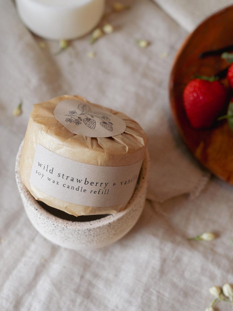Puede incluir: Recarga de vela de cera de soja envuelta en papel marr&oacute;n, etiquetada como "wild strawberry & vanilla". La vela se encuentra en un soporte de cer&aacute;mica blanca moteada. Una peque&ntilde;a ilustraci&oacute;n de fresas est&aacute; en la etiqueta.