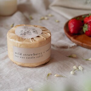 Puede incluir: Recarga de vela redonda de cera de soja, envuelta en papel marr&oacute;n, con la etiqueta "Wild Strawberry & Vanilla". La etiqueta presenta una ilustraci&oacute;n de fresa. Fresas frescas sobre una bandeja de madera. Sobre una superficie de lino neutra.
