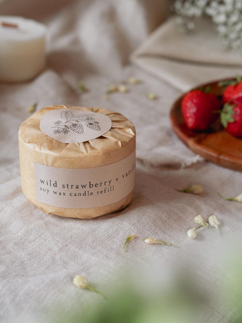 Puede incluir: Recarga de vela redonda de cera de soja, envuelta en papel marr&oacute;n, con la etiqueta "Wild Strawberry & Vanilla". La etiqueta presenta una ilustraci&oacute;n de fresa. Fresas frescas sobre una bandeja de madera. Sobre una superficie de lino neutra.