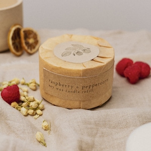 Może przedstawiać: Wypełnienie do świecy sojowej owinięte brązowym papierem z białą etykietą z napisem "raspberry & peppercorn soy wax candle refill". Świeca leży na beżowej powierzchni z białych kwiatów i czerwonych malin.