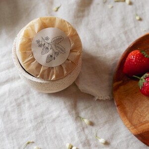 Puede incluir: Un peque&ntilde;o cuenco de cer&aacute;mica moteada con un art&iacute;culo envuelto, coronado con una pegatina con una ilustraci&oacute;n de fresa. Fresas frescas en un cuenco de madera a la derecha, con capullos de flores blancas esparcidos.