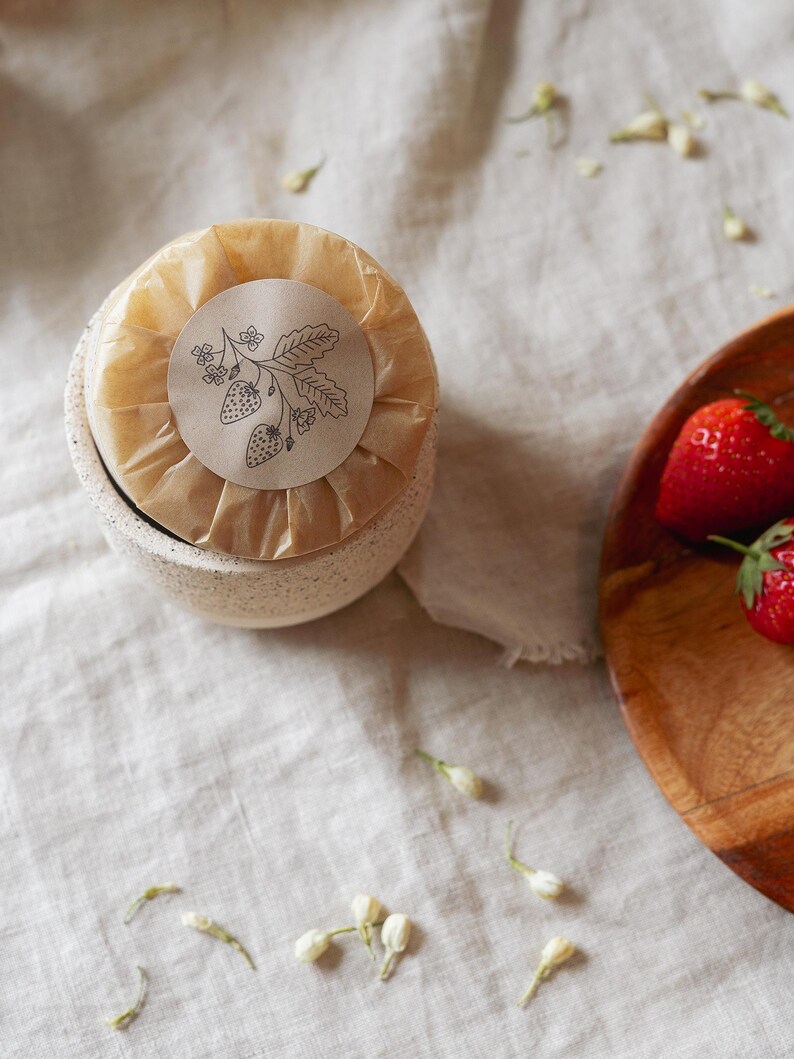 Puede incluir: Un peque&ntilde;o cuenco de cer&aacute;mica moteada con un art&iacute;culo envuelto, coronado con una pegatina con una ilustraci&oacute;n de fresa. Fresas frescas en un cuenco de madera a la derecha, con capullos de flores blancas esparcidos.