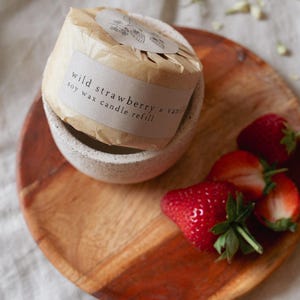 Puede incluir: Recarga de vela de cera de soja envuelta en papel marr&oacute;n, con la etiqueta "wild strawberry & vanilla". La vela est&aacute; en un cuenco de cer&aacute;mica moteada sobre una tabla de madera, con fresas frescas.