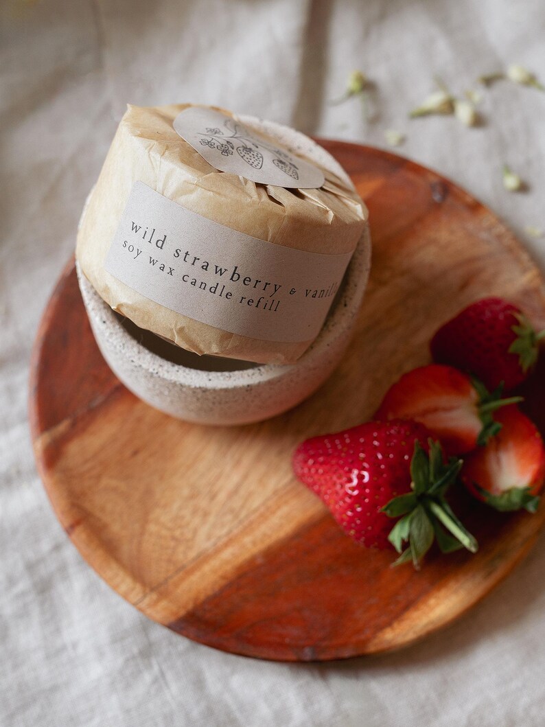 Puede incluir: Recarga de vela de cera de soja envuelta en papel marr&oacute;n, con la etiqueta "wild strawberry & vanilla". La vela est&aacute; en un cuenco de cer&aacute;mica moteada sobre una tabla de madera, con fresas frescas.