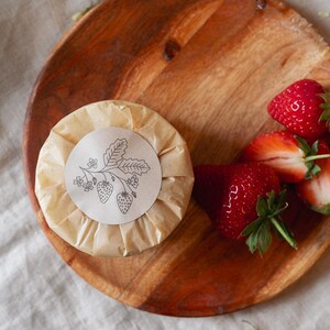Puede incluir: Un objeto redondo y envuelto con una ilustraci&oacute;n de fresa en una etiqueta de papel, sobre un plato de madera. Fresas frescas y maduras est&aacute;n dispuestas al lado. El plato es de madera marr&oacute;n claro.