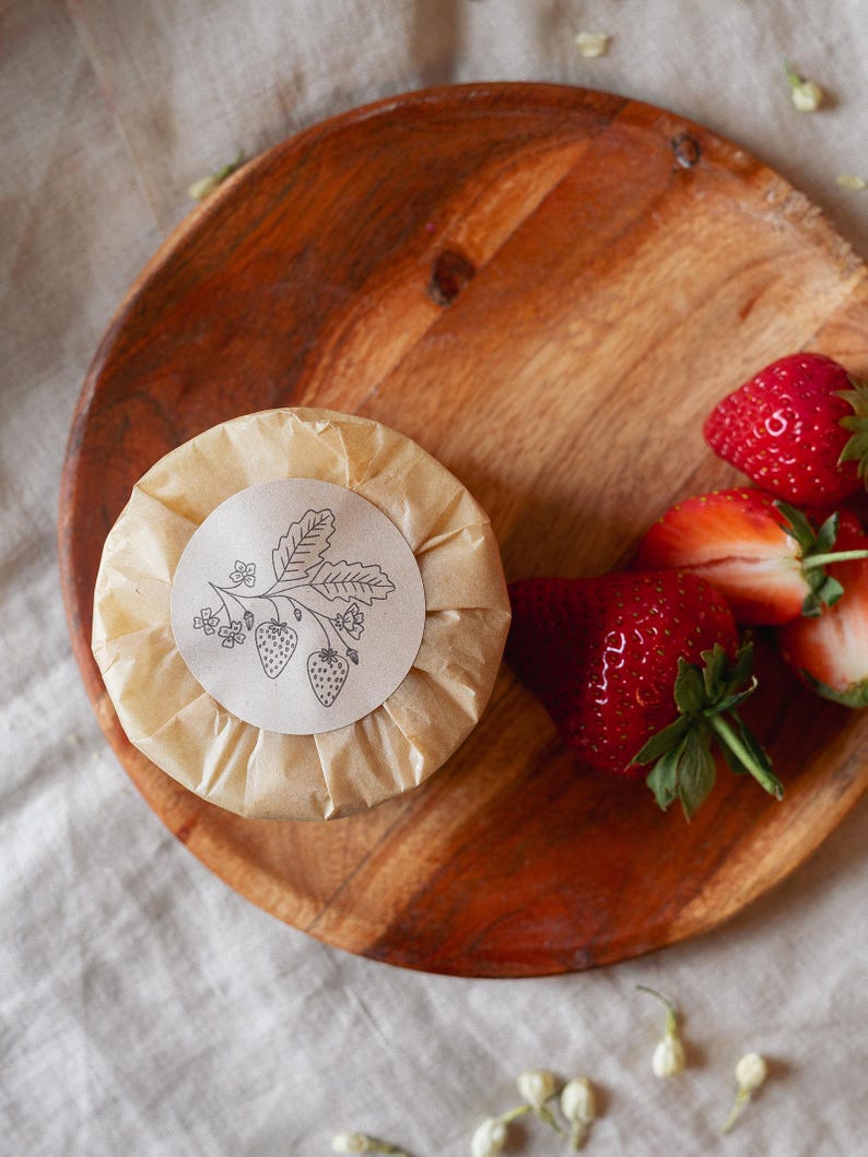 Puede incluir: Un objeto redondo y envuelto con una ilustraci&oacute;n de fresa en una etiqueta de papel, sobre un plato de madera. Fresas frescas y maduras est&aacute;n dispuestas al lado. El plato es de madera marr&oacute;n claro.