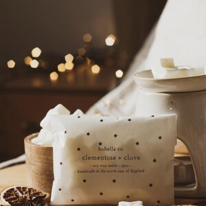 May include: A white package with black stars and the text "bobella co. clementine & clove - soy wax melts - 6pcs - handmade in the north east of England -" on it. The package is sitting on a wooden surface with a white wax melt in the shape of a star, a dried orange slice, and star anise.