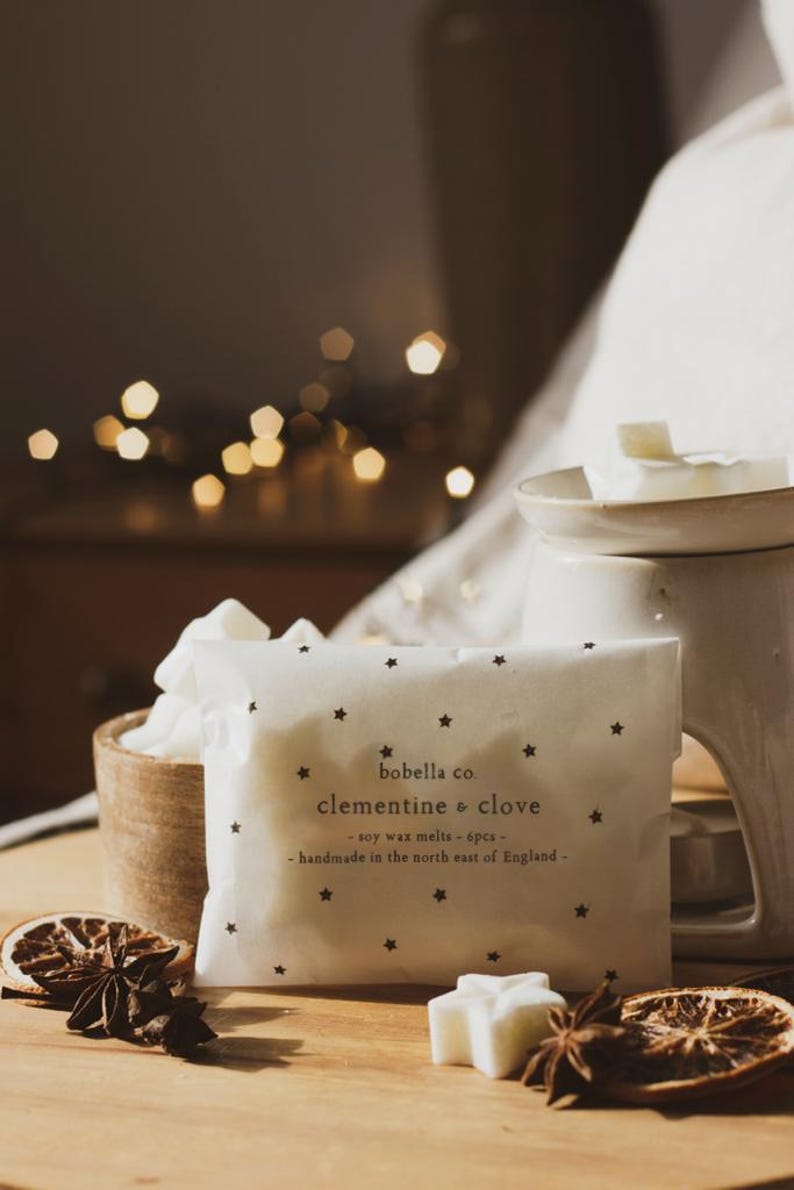 May include: A white package with black stars and the text "bobella co. clementine & clove - soy wax melts - 6pcs - handmade in the north east of England -" on it. The package is sitting on a wooden surface with a white wax melt in the shape of a star, a dried orange slice, and star anise.