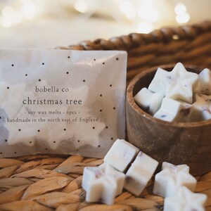May include: A package of six white soy wax melts shaped like stars, labeled "bobella co christmas tree - soy wax melts - 6pcs - handmade in the north east of England" in black text on a white background. The wax melts are in a small wooden bowl on a woven wicker surface.