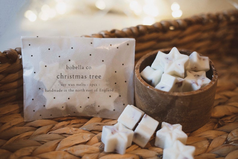 May include: A package of six white soy wax melts shaped like stars, labeled "bobella co christmas tree - soy wax melts - 6pcs - handmade in the north east of England" in black text on a white background. The wax melts are in a small wooden bowl on a woven wicker surface.