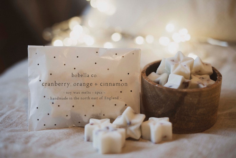 May include: A white bag with black polka dots and the text "bobella co cranberry, orange & cinnamon - soy wax melts - 6pcs - handmade in the north east of England -" printed on it. A wooden bowl filled with white star-shaped wax melts.