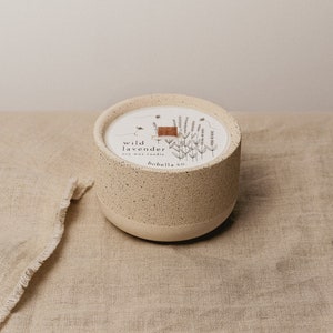 May include: A white ceramic candle jar with a speckled texture. The jar has a wooden lid and features a black and white illustration of lavender flowers and the text "wild lavender soy wax candle bobella co."