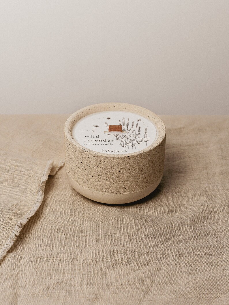 May include: A white ceramic candle jar with a speckled texture. The jar has a wooden lid and features a black and white illustration of lavender flowers and the text "wild lavender soy wax candle bobella co."