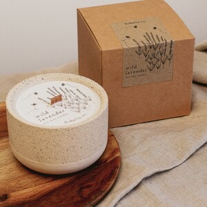 May include: A beige ceramic candle with a wooden wick and a label that reads "wild lavender soy wax candle" by "bobbella co.". The candle is sitting on a wooden coaster and a beige fabric background.
