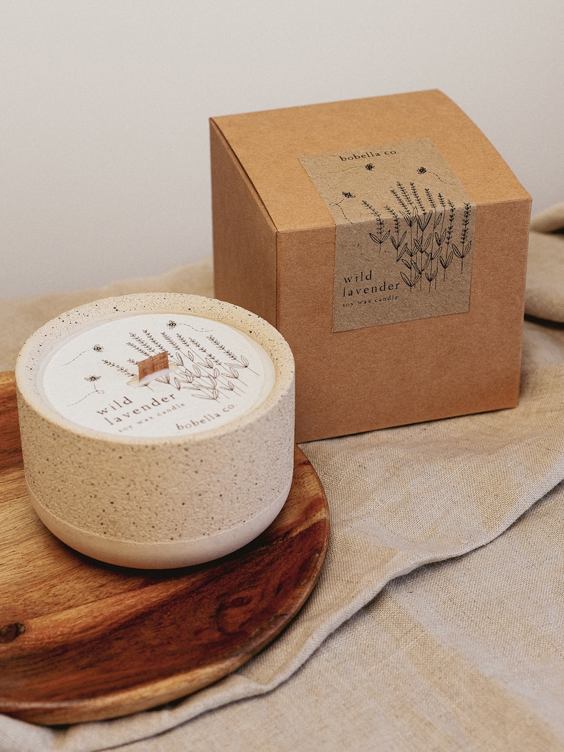 May include: A beige ceramic candle with a wooden wick and a label that reads "wild lavender soy wax candle" by "bobbella co.". The candle is sitting on a wooden coaster and a beige fabric background.