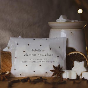 May include: A white package with black stars printed on it. The package contains soy wax melts in the scent of clementine and clove. The text on the package reads "bobella co clementine & clove - soy wax melts - 6pcs - handmade in the north east of England - "