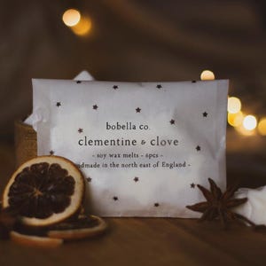 May include: A white package with a star pattern and the text "bobella co. clementine & clove - soy wax melts - 6pcs - handmade in the north east of England -" on it. The package is surrounded by dried orange slices, star anise, and white wax melts shaped like stars.