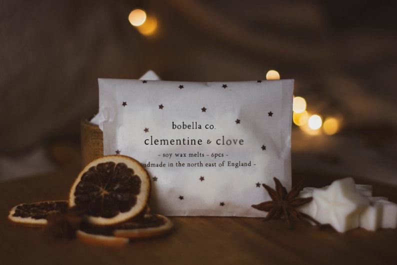 May include: A white package with a star pattern and the text "bobella co. clementine & clove - soy wax melts - 6pcs - handmade in the north east of England -" on it. The package is surrounded by dried orange slices, star anise, and white wax melts shaped like stars.