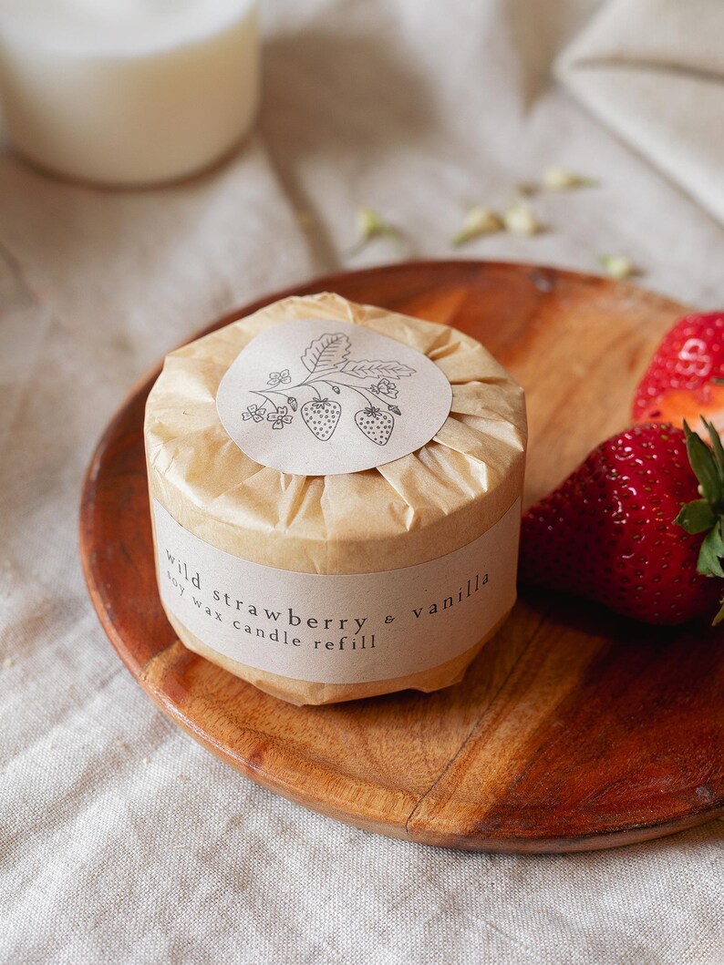Puede incluir: Recarga de vela redonda de cera de soja, envuelta en papel marr&oacute;n, con la inscripci&oacute;n "wild strawberry & vanilla". La etiqueta presenta una ilustraci&oacute;n de fresa. La vela est&aacute; sobre un plato de madera con fresas frescas.