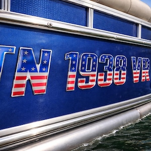 May include: Close-up of a blue boat hull with the letters "TN" and numbers "1938 VR" in a patriotic red, white, and blue design. The boat is on the water, with a blue fender visible.