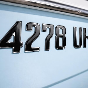 May include: Close-up of a boat's hull with the number "4278 UH" in glossy black lettering. The boat's hull is light blue, and a chrome rail is visible at the top. The numbers and letters have a reflective quality.