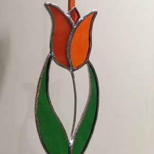 May include: A stained glass tulip ornament with orange petals and green leaves. The tulip is hanging from a gold ribbon.