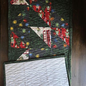 Könnte beinhalten: Ein gesteppter Weihnachtstischläufer mit dunkelgrünem Rand. Die Oberseite zeigt ein Patchwork-Design mit festlichen Mustern, darunter Lichterketten, rote Busse und Nussknacker. Die Unterseite ist weiß mit einem wellenförmigen Nahtmuster.