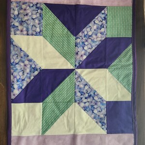 Può includere: Una trapunta rettangolare con bordo viola. La trapunta presenta un design patchwork con quadrati, triangoli e rettangoli nei toni del viola, bianco e verde. Alcune sezioni hanno un motivo floreale.