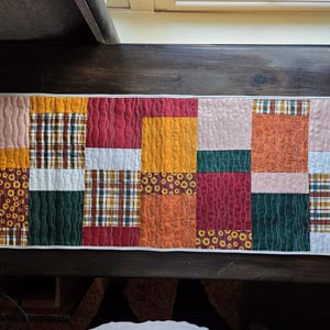 May include: A rectangular patchwork table runner with a variety of fabrics in warm tones. The design includes squares of floral, plaid, and solid textures in shades of red, orange, yellow, and green. The runner is approximately 12 inches wide.