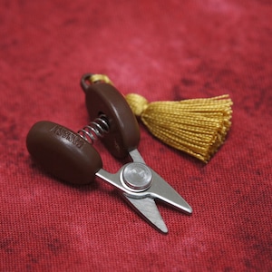 May include: Small brown and silver scissors with a gold tassel attached. The scissors have a spring mechanism and the handles are oval shaped. The scissors are on a red fabric background.