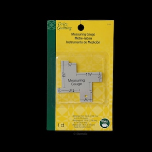 Puede incluir: Una galga de medición de plata con marcas en pulgadas y fracciones de pulgada. La galga está empaquetada en una tarjeta amarilla y verde con el texto "Dritz Quilting Measuring Gauge" y "1 ct".