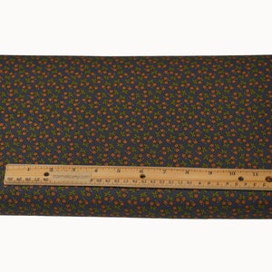 May include: A folded fabric with a dark gray background and a repeating pattern of small orange flowers and green leaves. A wooden ruler, marked in inches, rests on top of the fabric, showing measurements.