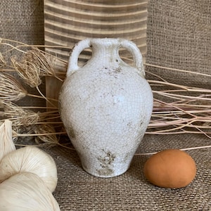 May include: A white ceramic vase with two handles and a cracked glaze. The vase has a rounded body and a narrow neck. An egg and garlic bulbs are in the foreground. The background includes dried plants and a wooden board.