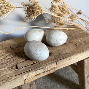 May include: Three smooth, oval-shaped stones with a mottled light grey and beige surface texture. The stones are arranged in a cluster, displaying natural variations in colour and tone across their rounded, solid forms.