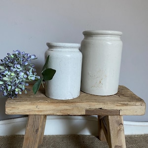 May include: Two cylindrical off-white ceramic crocks with speckled textures and a smooth finish. The crocks are positioned next to a cluster of purple and blue hydrangea flowers, featuring rounded rims and straight sides.