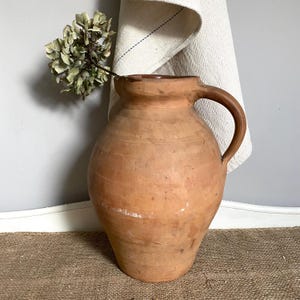 May include: A large, round, terracotta pitcher with a single curved handle and a wide mouth. The surface is matte, earthy brown, and features subtle horizontal ridges. A single dried, pale green hydrangea flower stem is placed inside the pitcher.