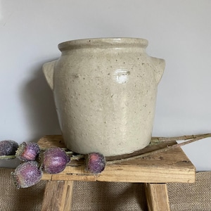 May include: A light beige ceramic crock with a speckled finish, a wide rim, and two small side handles. The vessel has a cylindrical body with a slightly tapered base and a smooth, glazed texture, showcasing natural variations in the clay material.